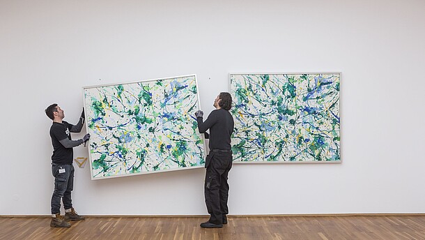 Two people are hanging a painting on a wall next to a painting that already is hung up.