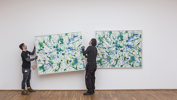 Two people are hanging a painting on a wall next to a painting that already is hung up. 