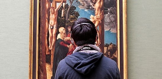 A person with their back to the camera is wearing headphones and is looking at a painting in front of them. 