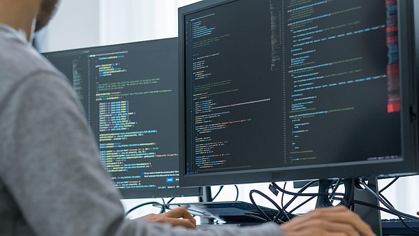 The focus is on two computer screens with code that someone is writing. Only the hands and the side of the shoulder and parts of the face is visible. 