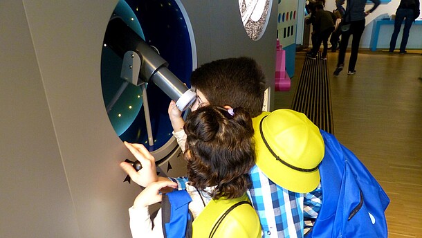 Two children wearing yellow construction hats are taking turns to look into a telescope.