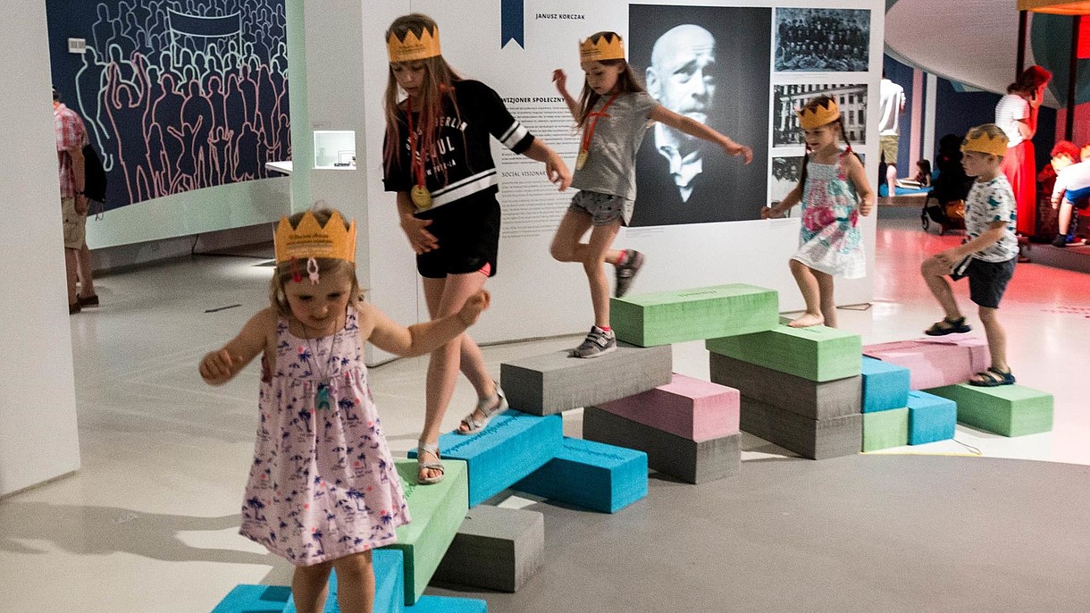  Children with paper crowns on their heads are balancing on stones inside an exhibition space.