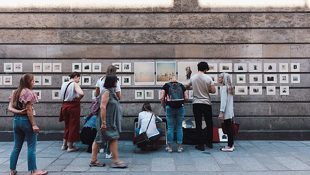  People walk on a street where several pictures have been hung on a wall creating a kind of outside gallery. The people look at the pictures as they walk by.