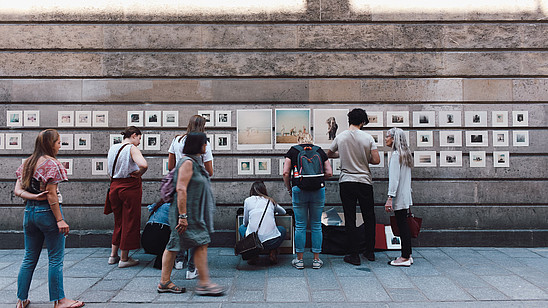 People walk on a street where several pictures have been hung on a wall creating a kind of outside gallery. The people look at the pictures as they walk by.
