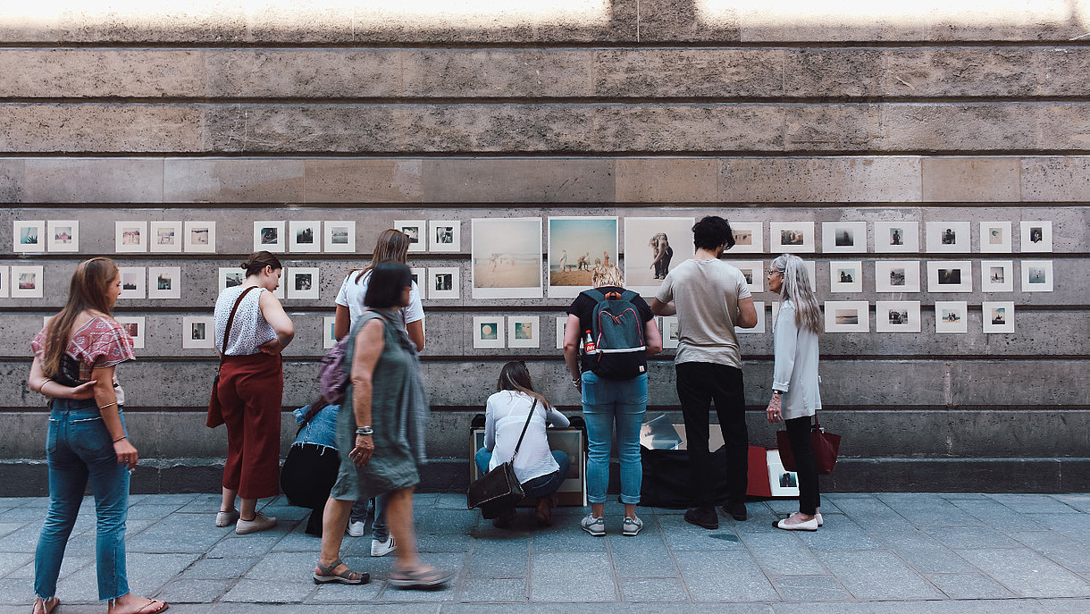 © Image: Tetiana Shevereva People walk on a street where several pictures have been hung on a wall creating a kind of outside gallery. The people look at the pictures as they walk by.