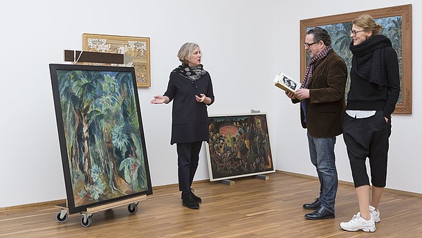  A woman is showing two people a painting that is standing on the ground. The man is reading about the painting in a book.