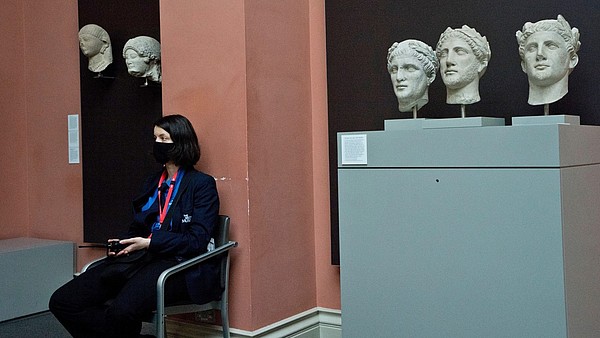 A security officer, wearing a face mask, sits next to ancient stone busts inside a museum space