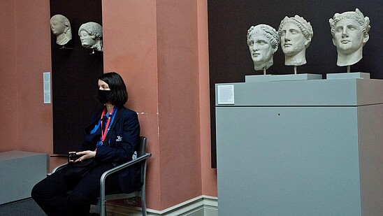 A security officer, wearing a face mask, sits next to ancient stone busts inside a museum space