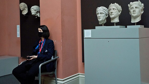 A security officer, wearing a face mask, sits next to ancient stone busts inside a museum space