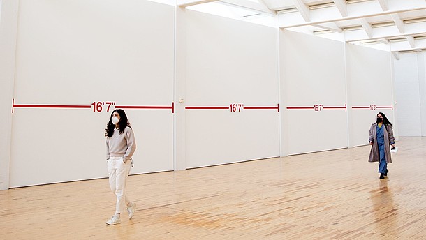 Two people wearing face masks walk through an exhibition hall. There is plenty of space between them and the walls are covered with distance markings.  