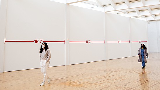 Two people wearing face masks walk through an exhibition hall. There is plenty of space between them and the walls are covered with distance markings.