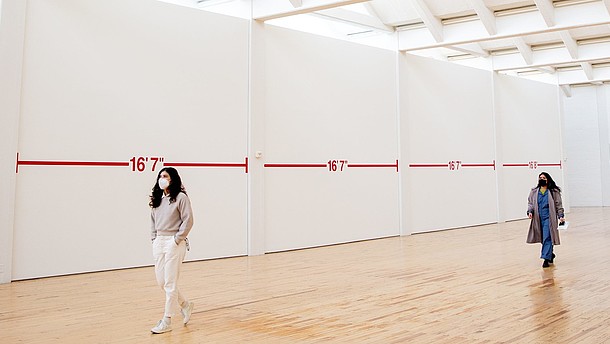  Two people wearing face masks walk through an exhibition hall. There is plenty of space between them and the walls are covered with distance markings.