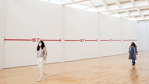Two people wearing face masks walk through an exhibition hall. There is plenty of space between them and the walls are covered with distance markings.