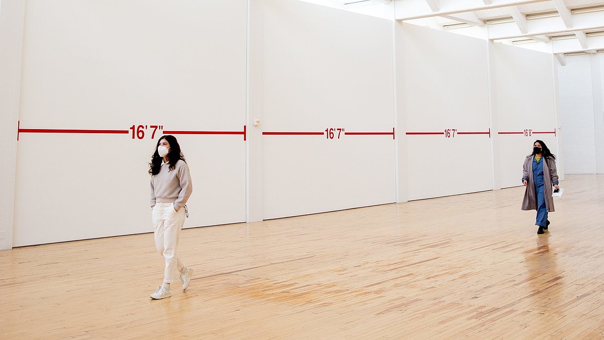  Two people wearing face masks walk through an exhibition hall. There is plenty of space between them and the walls are covered with distance markings.