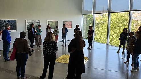 Group of people in an exhibition hall with pictures on the wall and an art piece on the floor in the middle of the room. A guide is talking to the people.  