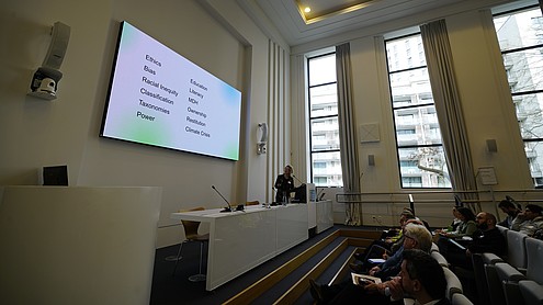 One person is standing behind a podium speaking in front of a seated group of people. She looks back on a projected image that reads “Ethics, Bias, Racial Inequality, Classification, Taxonomies, Power, Education, Literacy, MDH, Ownership, Restitution, Climate Crisis”.   © Image: Peter Vand der Plaetsen